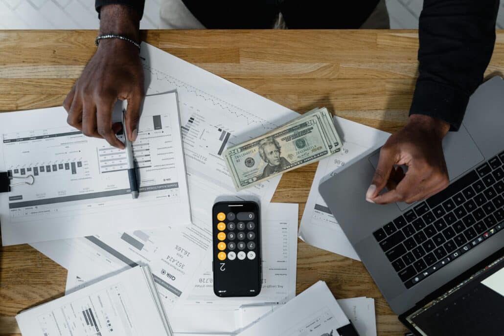 person surrounded by papers with graphs on them while at a desk with a calculator, cash, and an open laptop on it