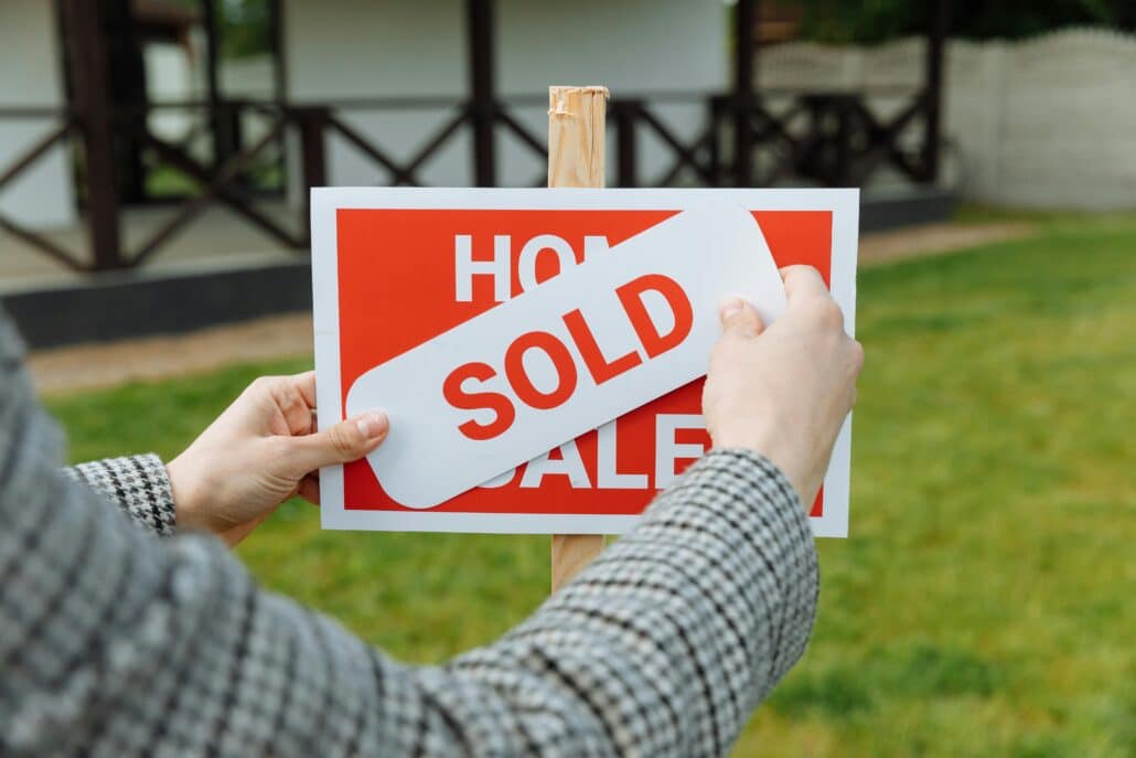 a red and white "house for sale" sign in a front lawn being covered by a white sticker that spells out "sold"