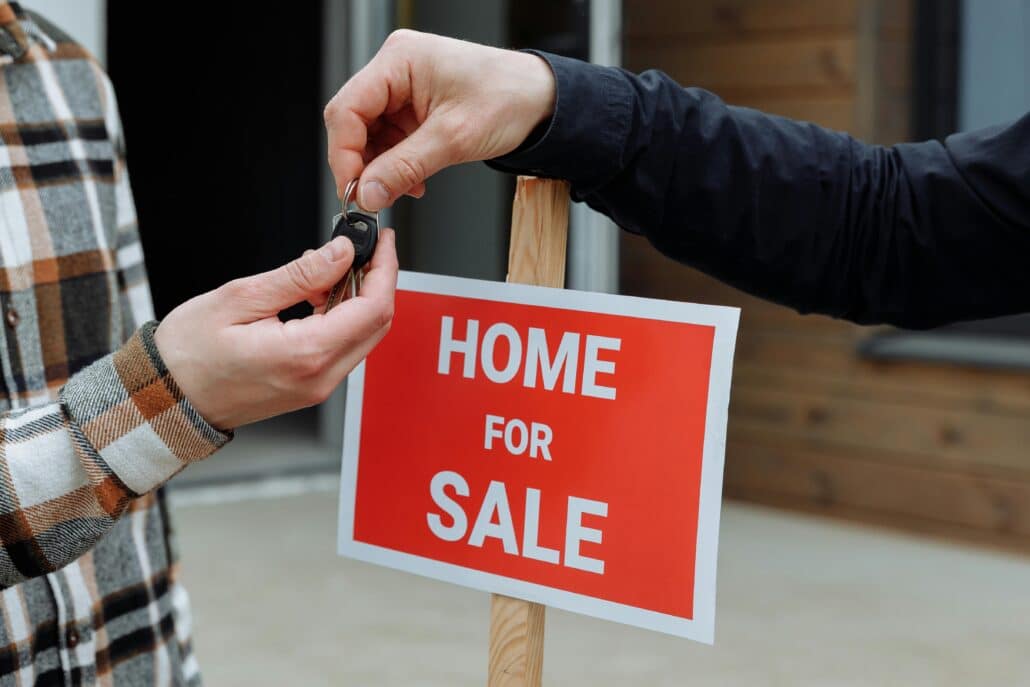 someone handing over a set of keys in front of a "home for sale" sign