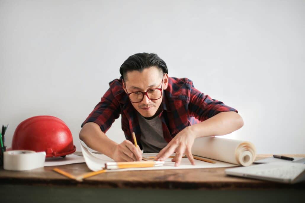 man drawing on building plans with a red hard hat next to him